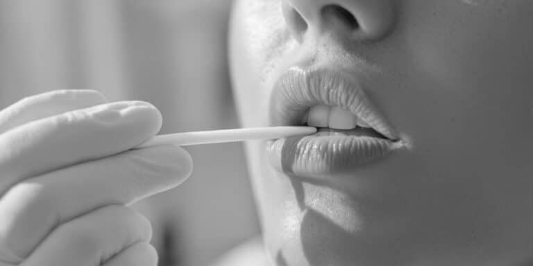 Close-up of a person holding a DNA testing swab near their lips, which are slightly parted. The gloved hand suggests a careful, possibly medical or hygienic procedure. The image is in black and white, capturing the precision of this mouth swab moment.